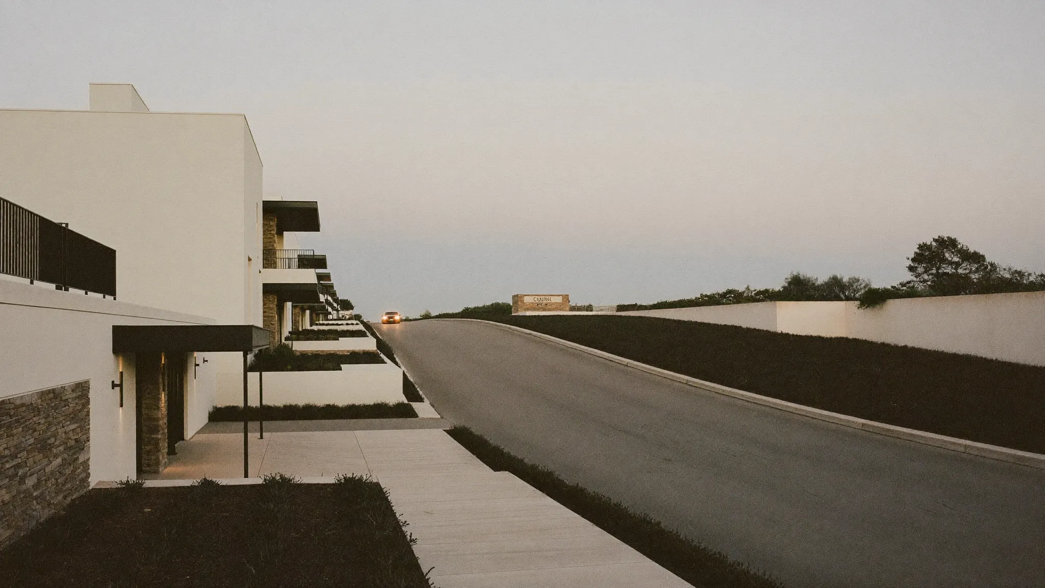 An apartment community entrance drive at dusk, a single car approaching with its headlights on
